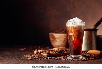 Ice Coffee With Cream Being Poured Into It Showing The Texture And Refreshing Look Of The Drink. Frozen Glass And Coffee Beans On A Old Brown Table With Kitchen Utensils. Copy Space.