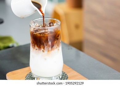 Ice Coffee Latte On A Wooden Table With Black Coffee Being Poured Into It Showing The Texture And Refreshing Look Of The Drink. Iced Coffee In A Tall Glass With Coffee Poured Over Milk. 