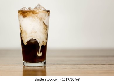 Ice Coffee On A Wood Table With Cream Being Poured Into It Showing The Texture And Refreshing Look Of The Drink, With A Clean Background.