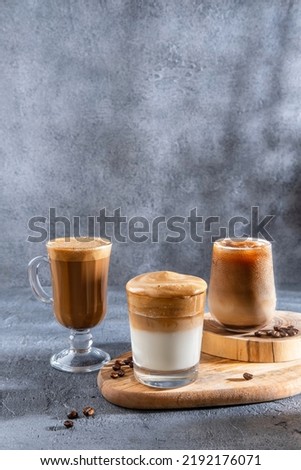 Ice coffee in a tall glass with cream poured over and coffee beans. Set with different types of coffee drinks on a dark table.