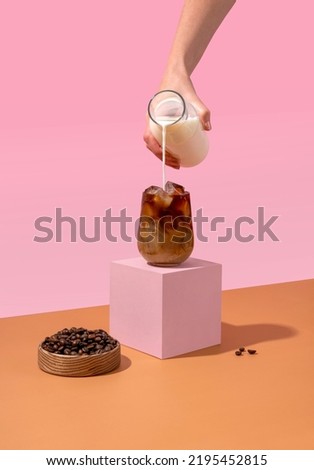Ice coffee in a tall glass with cream poured over and coffee beans. A woman's hand pours cream into a glass of coffee.
