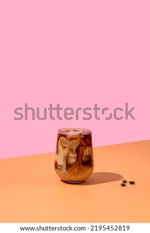 Ice coffee in a tall glass with cream poured over and coffee beans on pink background. 