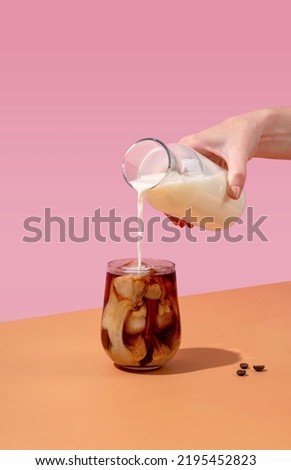Ice coffee in a tall glass with cream poured over and coffee beans. A woman's hand pours cream into a glass of coffee.