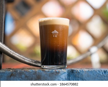 Ice Cold Nitro Dispensed Glass Of Dark Beer With Foam Outside On A Sunny Day. Stout Beer Freshly Poured With Big Head On Blue Shelf. Cup Of Beer In Front Of Lattice. 