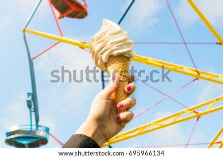 Ice cream in amusement park on a sunny summer day. Girl or young woman holding a cone of soft ice with a colorful theme park ride in the background.