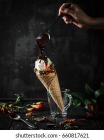 Ice Cream Cone In A Glass Cup With Woman's Hand Holding A Spoon Of Melted Chocolate Pouring On Ice Cream On A Black Background, Low Light