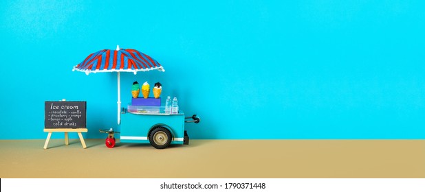 Ice cream toy cart with blue red umbrella. Assortment of ice cream menu black chalkboard. Summer vacation concept. Blue sandy beach background. copy space.