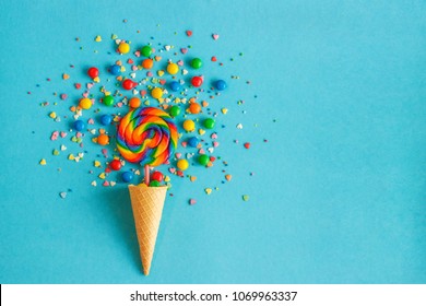 Ice cream waffle cone with colorful lollipop on stick, scattering of multicolored sweets and confectionery topping. Blue background