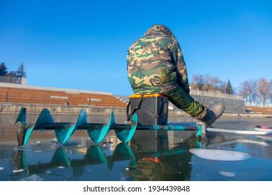 Ice Fishing Auger And Fisherman Sitting In The Background On The Ice