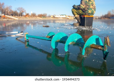 Ice Fishing Auger And Fisherman Sitting In The Background On The Ice