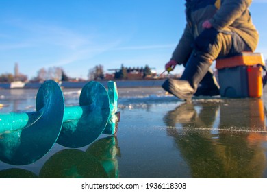 Ice Fishing Auger And Fisherman Sitting In The Background On The Ice