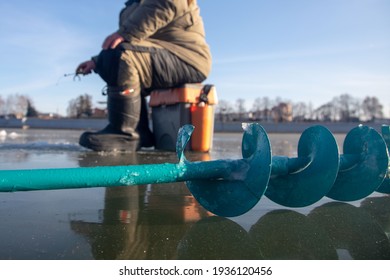 Ice Fishing Auger And Fisherman Sitting In The Background On The Ice