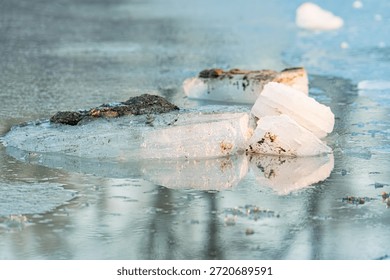 Ice patch floating on river, rough and irregular with dark specks Clear undisturbed water reflection mirroring ice shape Light source natural, casting shadow beneath ice Muted grays dominating sce