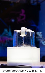 An Ice Sculpture With Two Ice Blocks And A Candle In The Sculpture On A Table With A White Material With Black Letters With A Blurry Background .