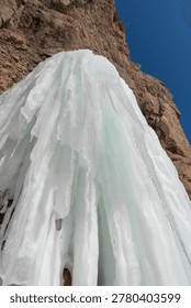 Ice structure flows down a rocky cliff, illuminated by sunlight against a bright blue sky, highlighting the beauty of natural ice formations