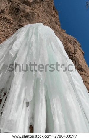 Ice structure flows down a rocky cliff, illuminated by sunlight against a bright blue sky, highlighting the beauty of natural ice formations