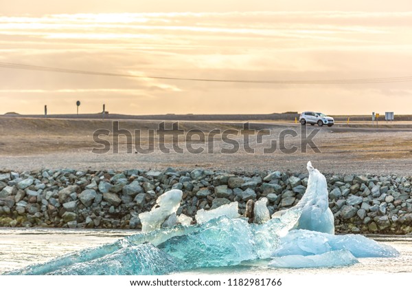 Iceberg floating in a river with a SUV\
car parked in the background, sunset colors in\
Iceland