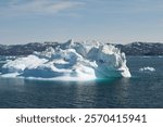 Iceberg floats in the deep blue waters of the icefjord near the Eqip Sermia Glacier in Greenland, with distant snow-capped mountains under a clear sky