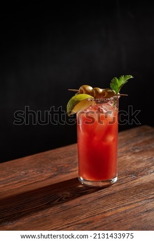 Iced bloody Mary (Caesar) cocktail with olives, parsley and lime piece in a glass, on a wooden bar and dark background. Vertical photo with shallow depth of field and copyspace.   