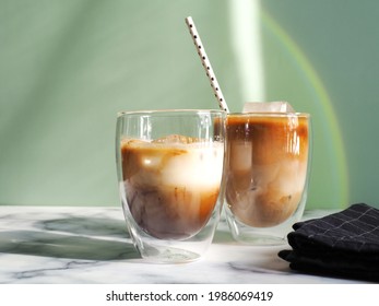 Iced Caramel Latte Coffee In Double Wall Insulated Glasses With Fancy Eco Friendly Paper Straw On White Marble Background And Green Wall With Rainbow Light Reflection. Refreshing, Enjoy Summer Drinks.