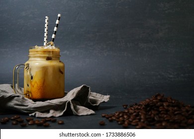 Iced Caramel Latte Coffee In A Glass Jar Drinking Mug With Handle And Fancy Paper Straws On Dark Background With Napkin And Coffee Beans. Refreshing And Enjoy Summer Drink Theme. (space For Text)