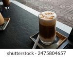 Iced caramel latte topped with caramel sauce, a refreshing and sweet coffee drink. Ceramic coasters. On a black table. Work area in a cafe. There is a notebook next to the glass of water.