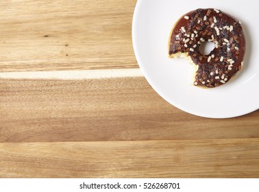 An Iced Chocolate Sprinkle Ring Donut With Bite Mark, On A Wooden Kitchen Counter Top Background With Empty Space At Side