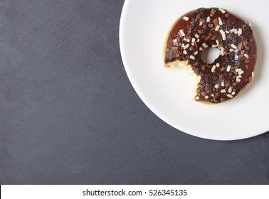 An Iced Chocolate Sprinkle Ring Donut With Bite Mark, On A Rustic Slate Background With Empty Space At Side