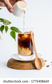 
Iced coffee with cream in a tall glass. Drink composition on a white background. Hand pouring cream into coffee. Summer refreshing beverage