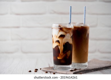 Iced coffee latte cappuccino in a tall glass with cream or milk and coffee beans and straws on a white wood background. Cold tasty summer refreshment beverage concept. Selective focus, copyspace.