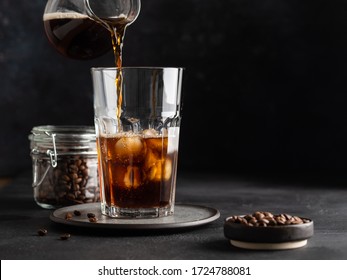 Iced coffee, process of making. Pouring coffee into glass with ice cubes. Cold and refreshing beverage, soft drink. Dark background. Copy space.