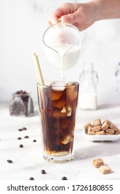 Iced coffee in a tall glass with cream being poured into it showing the texture of the drink. Cold coffee with ice cubes.