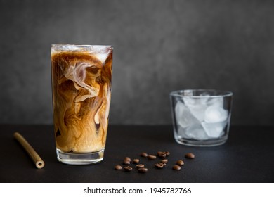 Iced coffee in tall glass with cream, container with ice, cocktail straw and coffee beans on dark background with copy space. Refreshing drink.