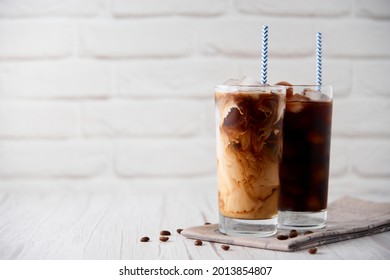 Iced coffee in a tall glass with cream on a white wood background. Cold tasty summer refreshment beverage concept. Selective focus, copyspace.