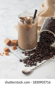 Iced Cold Coffee With Milk And Glass Container Of Beans And Spoon With Salted Caramel On Marbel Board And Light Table Background.