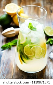 Iced Lemonade Pitcher, Wooden Juicer, Cold Citrus Infused Water Beverage With Lemon & Lime Slices, Mint Leaves On Brown Grunged Wood Textured Table Sunny Day, Grunged Background. Close Up, Copy Space.