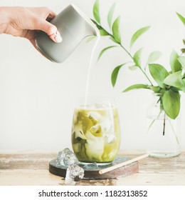 Iced Matcha Latte Drink In Glass On Board With Coconut Milk Pouring From Pitcher By Hand, White Wall And Plant Branches At Background, Copy Space, Square Crop. Summer Refreshing Beverage Cold Drink