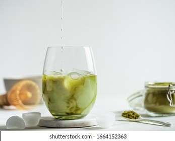 Iced Matcha Latte Tea with pouring milk drops into tumbler glass. Matcha latte and ingredients on white marble background. Copy space for txt or design - Shutterstock ID 1664152363