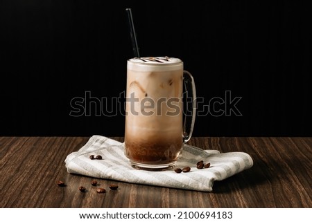 Iced mocha coffee with cream in a tall glass and coffee beans, portafilter, tamper and milk jug on dark wooden background. Cold summer drink.