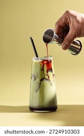 Iced raspberry matcha drink with red syrup being poured from a jigger into a clear glass. Colourful layered beverage with ice on a clean, minimal studio background, hands only.