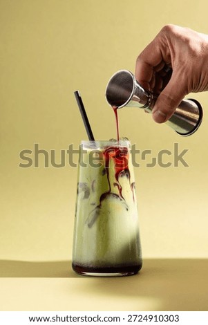 Iced raspberry matcha drink with red syrup being poured from a jigger into a clear glass. Colourful layered beverage with ice on a clean, minimal studio background, hands only.