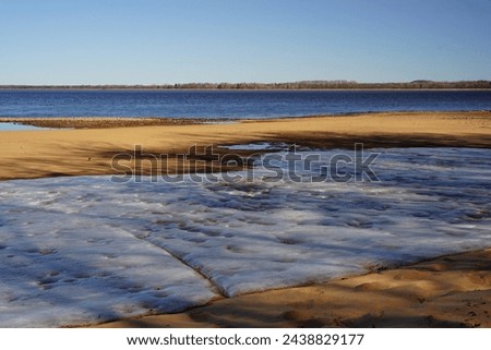 Iced snow covers a sandy beach on low tide lake. 
