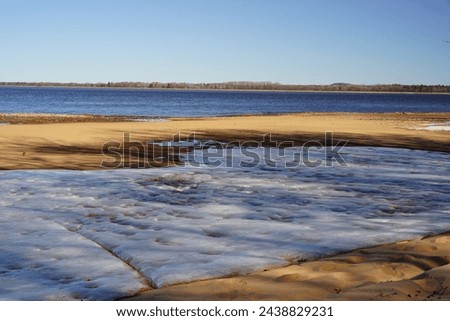 Iced snow covers a sandy beach on low tide lake. 