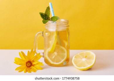 Iced tea with lemon slices and mint, glass pitcher with citrous refreshing summer cocktail and drinking straw on table decorated with flower and half of lemon.