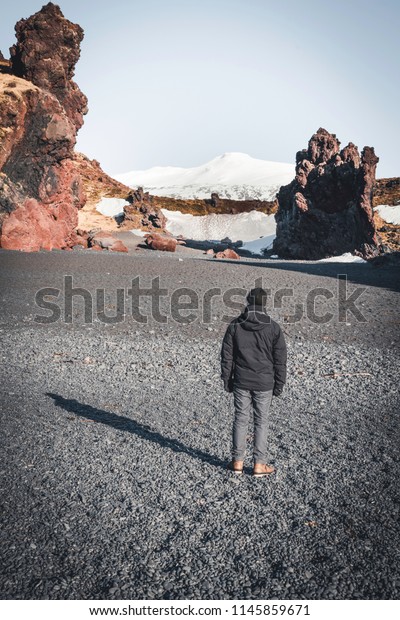 Iceland Black Beach Sand young man\
tourist traveller with snaefellsnesjokull mountain snow blue sky in\
Background. Western side if the\
country.