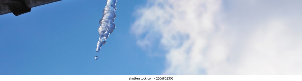 Icicle against blue sky and white cloud. Drop of melted snow falls down. Close up. Icicle hangs from rain gutter on roof. Banner or headline about end of winter or beginning of spring. Thaw. Macro
