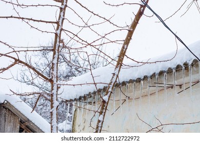 Icicles Hang From The Roof Of A Village House On A Winter Day. Snow Melting.