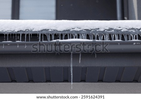 Icicles hanging from a snow-covered eave and black gutter on a modern house during winter, showing frozen water runoff. High quality photo