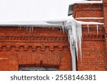 Icicles on gutter and roof. Huge Icicles hanging down from drain pipe of the red building. Icy facade of building, dangerous icicles melt on rooftop, risk of injury. Ice hazard