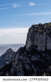 Iconic cross atop a dramatic limestone cliff in the Montserrat mountain range, Catalonia, Spain, overlooking a vast valley and distant mountains under a clear sky.